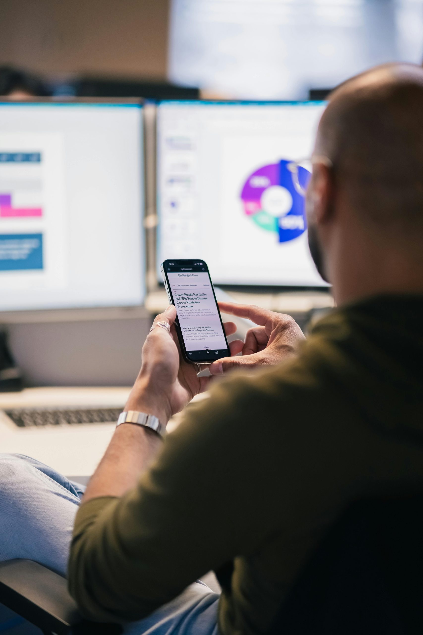 A man views his smartphone with an article titled "Ready to Qualify" while sitting at a desk with two computer monitors displaying data and charts in the background.