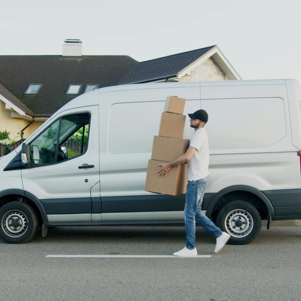 A delivery driver wearing a cap and jeans carries a stack of cardboard boxes from a silver cargo van parked on a suburban street.