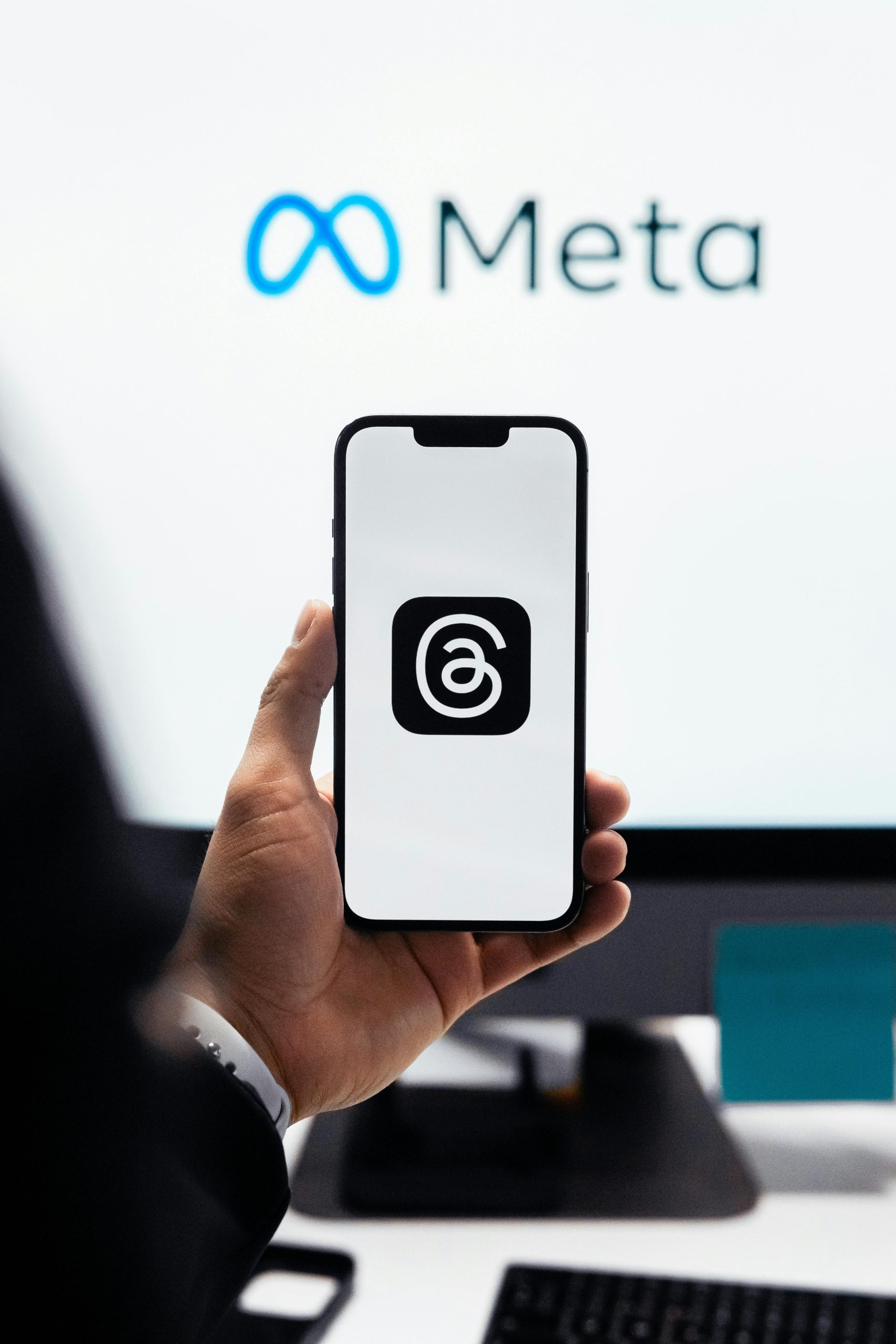 A hand holds a smartphone displaying the Threads app icon, with the Meta logo (infinity symbol and text) visible on a monitor screen in the background.