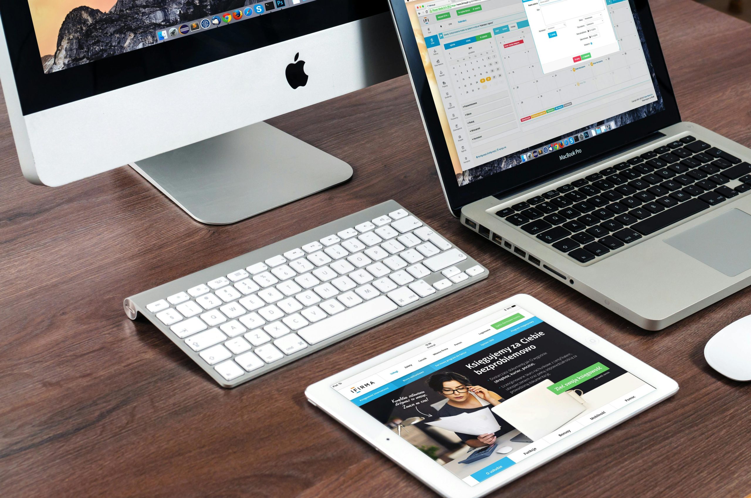 An overhead view of a desktop computer, laptop, wireless keyboard, and tablet displaying a website, all on a wooden desk.